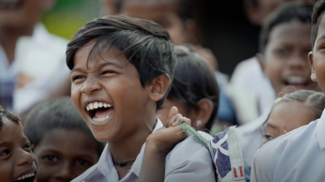 A young boy in a school uniform laughs joyfully, surrounded by other smiling children. The group appears happy and engaged, creating a lively, cheerful scene.